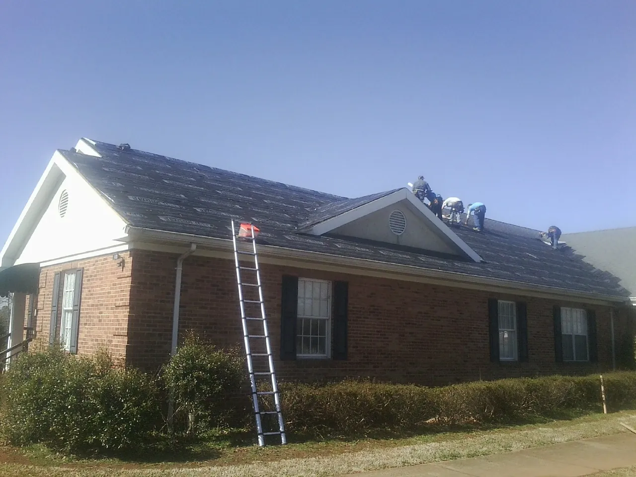Crew tearing shingles off a brick ranch roof with a ladder set — Emergency Roof Repair work by our crew in Cassatt