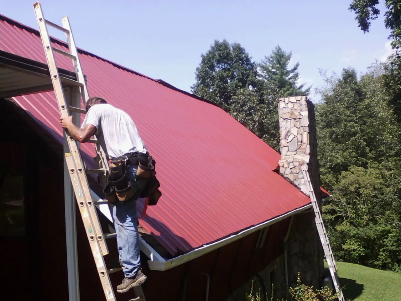 Roofer on a ladder beside a red metal roof and stone chimney — Soffit & Fascia Repair by Roofing Brothers in Cassatt